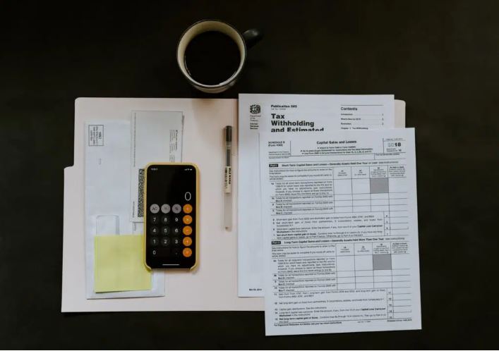 IRS tax forms, IPhone calculator,and coffee cup on a table to signify paying taxes and helping Self-Directed IRA account holders avoid extra taxes and penalties.