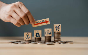 Stack of coins with blocks on top with images of a dollar sign, oil tank, dollar bill, shopping cart, and a red arrow to show increasing inflation.