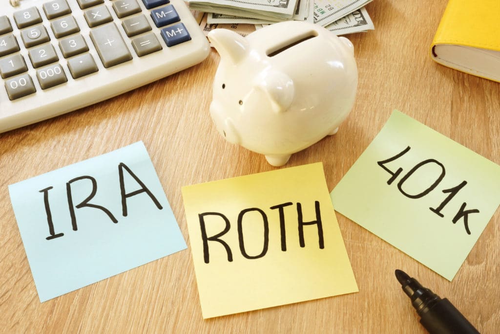 Piggy bank, calculator, and sticky notes with “IRA,” “Roth,” and “401k” written on them to display contributing funds to a retirement account.