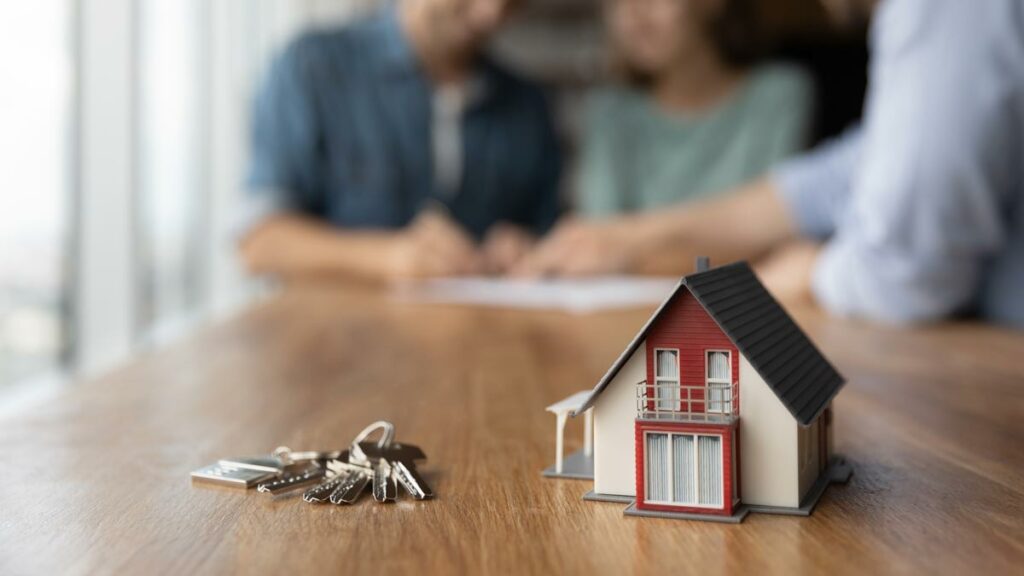 A miniature house and house keys sitting on a table with a couple and a financial advisor in the background signing non-recourse loan documents.