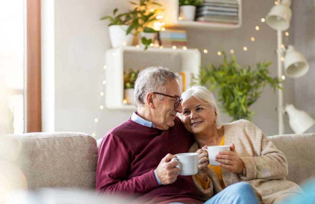 Retired couple on the couch drinking tea and smiling as they can enjoy what they planned for in retirement.