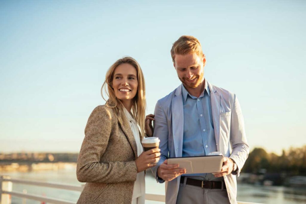 Young, successful Self-Directed IRA account holders standing with a sunset background, holding coffee, and looking at a tablet with their self-directed investing strategy on it.