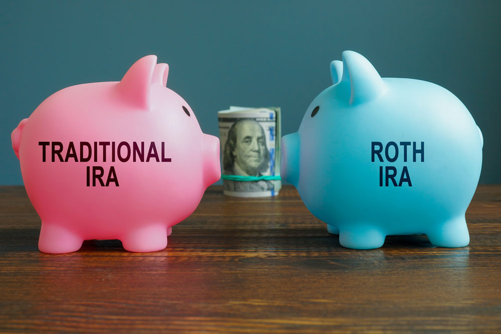 Two piggy banks, one pink that says, “Traditional IRA” and one blue that says “Roth IRA” facing each other with a stack of money in the middle to show the difference between retirement accounts.  