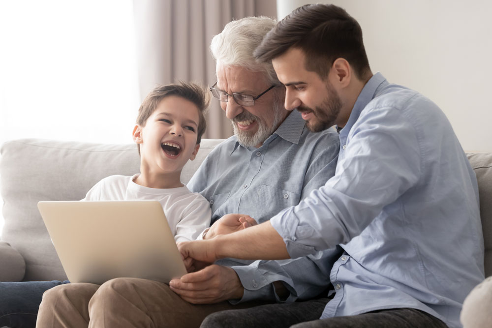Three generations (grandfather, son, and grandson) smiling and laughing at a computer screen as the grandfather plans his Self-Directed IRA beneficiaries.