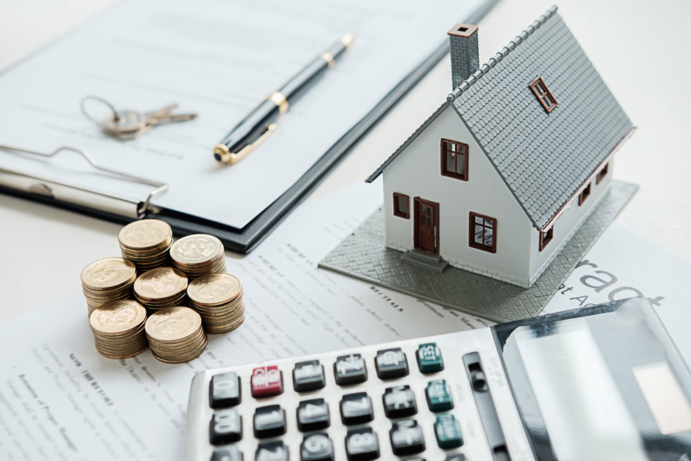 Gold coins, a clipboard, a small model house, and a calculator arranged on a table, showing an investment in real estate with a Self-Directed IRA.