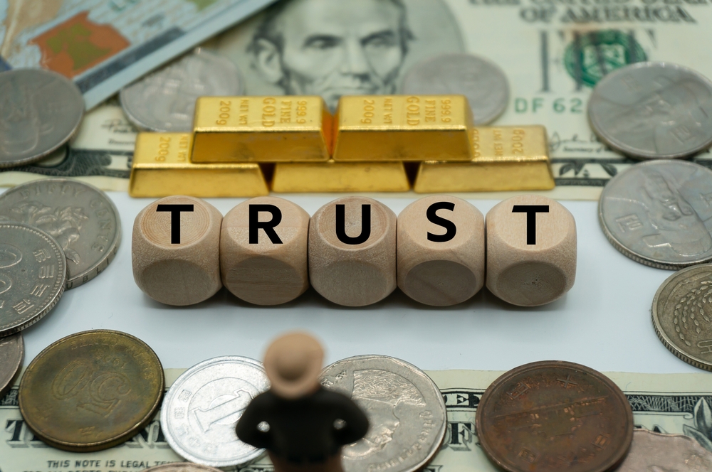 The word "trust" spelled out with wooden Scrabble cubes on a table surrounded by silver coins, $5 bills, and small golden bars