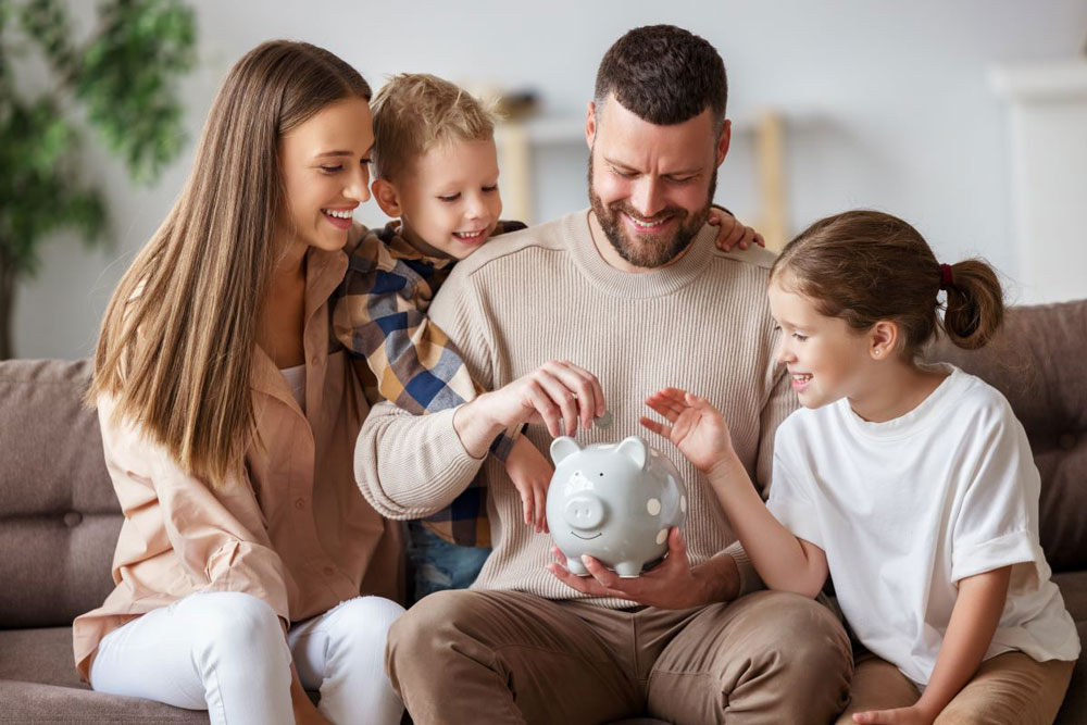 A family gathered on the couch, smiling, and putting coins in a piggy bank to display fun ways the entire family can save.  