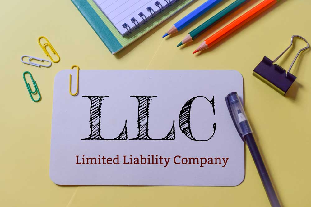 Purple notecard that says “LLC Limited Liability Company” on a desk with colored pencils, a notebook, and paper clips.