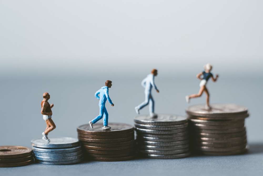Running figurines sprint across increasing stacks of coins, indicating that not only does fitness go hand in hand with Self-Directed IRA investing, but planning for retirement leads to greater savings as the years progress.