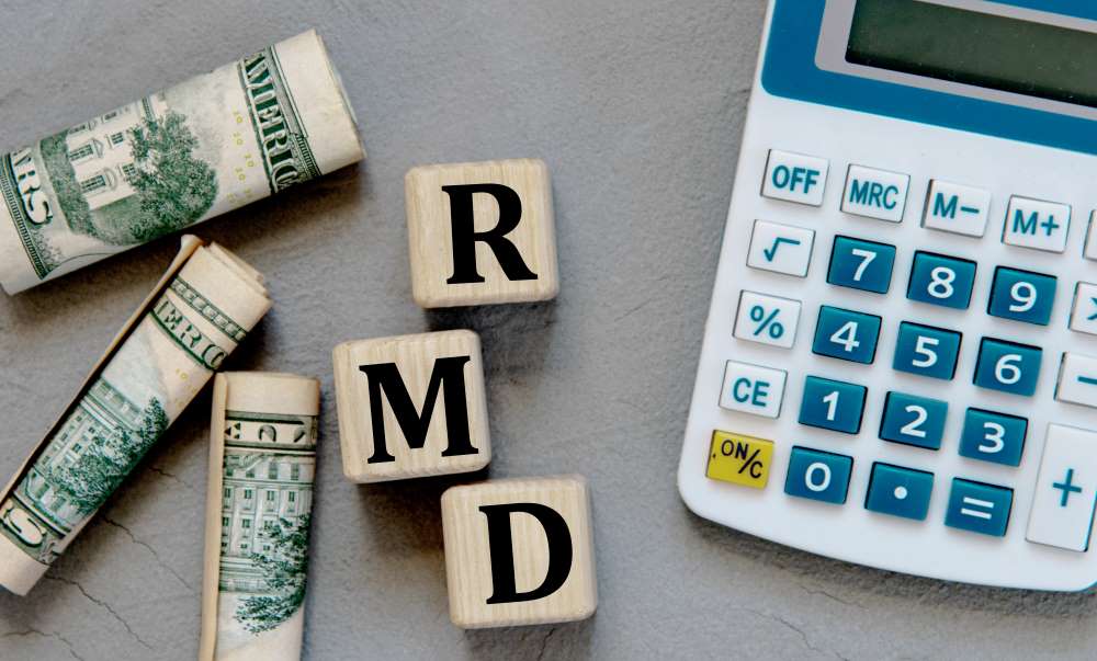 A calculator rolled up dollar bills, and blocks depicting the phrase ‘RMD,’ showcasing how it’s best to come up with a plan to have your rental income cover your RMDs.