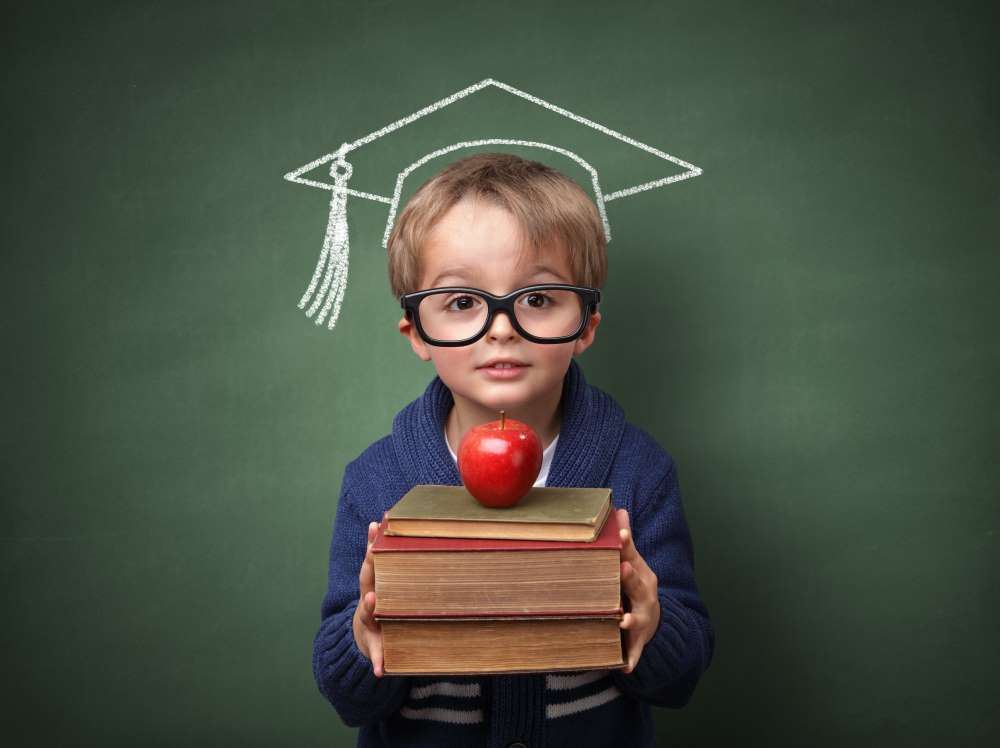A young boy stands with a pile of books and a graduation cap, indicating that he is actively receiving a financial education and is ready for the establishment of his Custodial Self-Directed IRA.