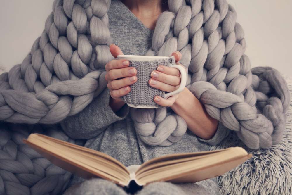 A woman drinks tea and reads a book, as she cozies up under her Self-Directed IRA BLANKET. 