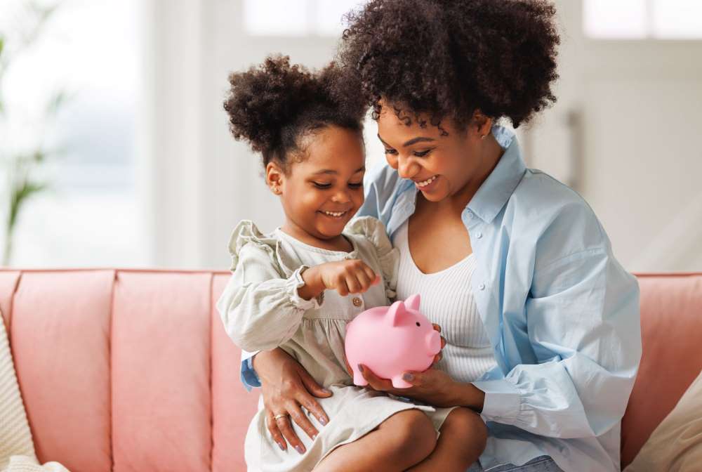 A mother and daughter discuss saving for the future as they place coins in a piggy bank, in preparation for her Custodial Self-Directed IRA.