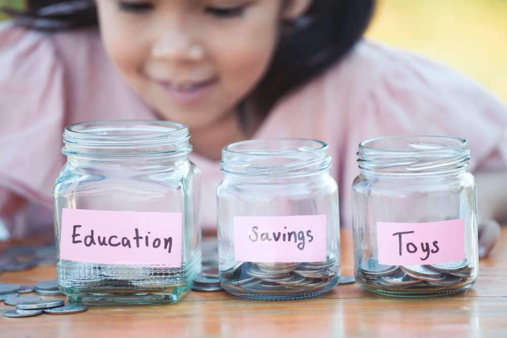 A child divides her money into three jars labeled “Education,” “Savings,” and “Toys” after she has learned about financial literacy from her parents.