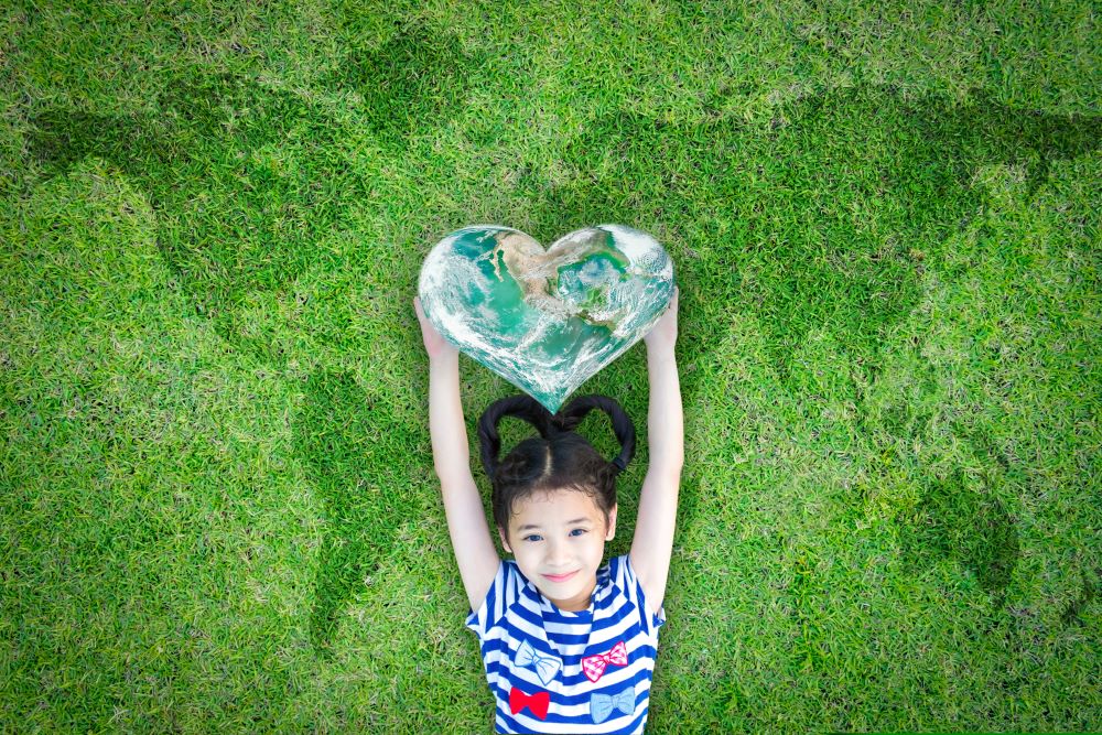 A girl lies on the grass, holding the world in the shape of a heart, promoting Random Acts of Kindness Day. 