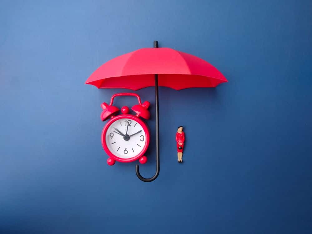 A red umbrella hangs over a clock and a figurine, showcasing how preparedness and an emergency fund is a crucial element to financial wellness.