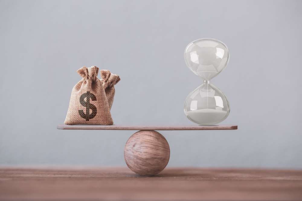 A seesaw with a money bag on one end, and an hourglass on the other, depicting that time and money are one in the same.