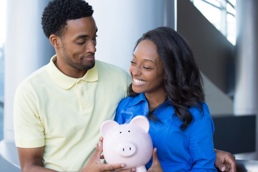 A happy couple nestles with their piggy bank, excited that their retirement savings will be developing tax-free in their Self-Directed Roth IRA.