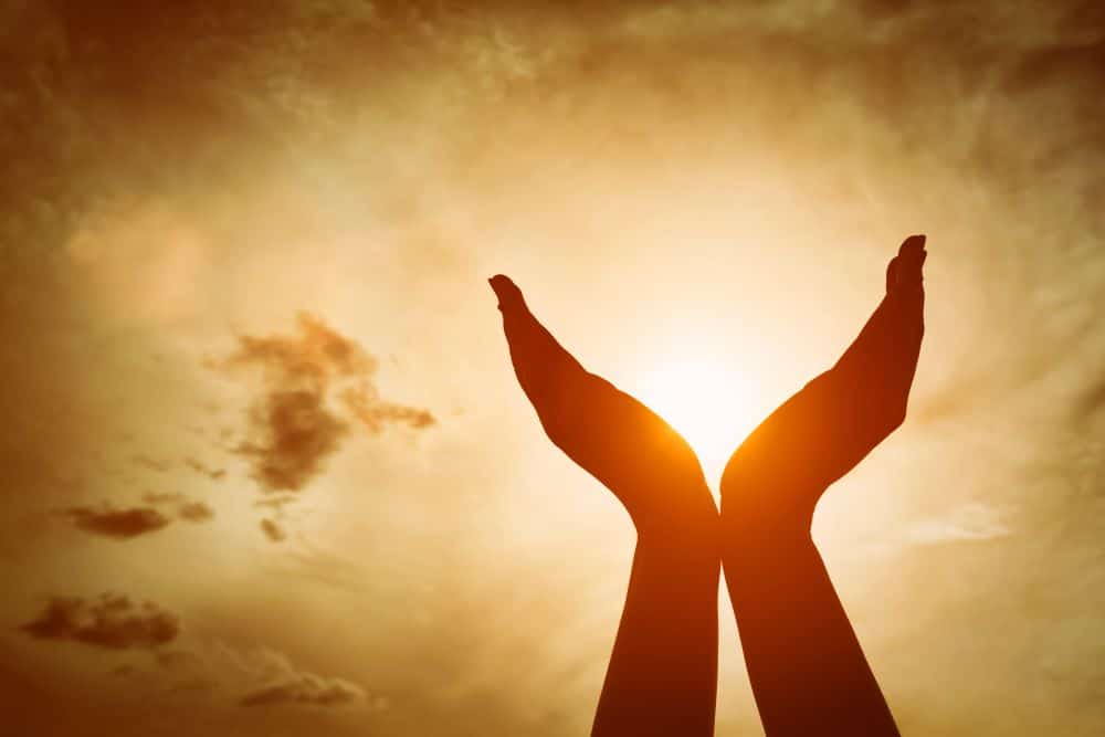 Hands stretch up towards a beautifully lit sky, indicating hope and the rewards received from pursuing self-love and financial wellness. 