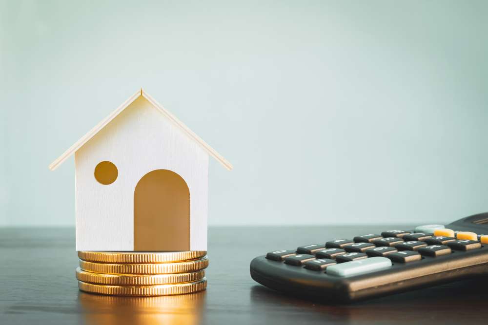A house sits on top of a stack of coins beside a calculator, depicting that real estate is an asset that tends to appreciate over time.