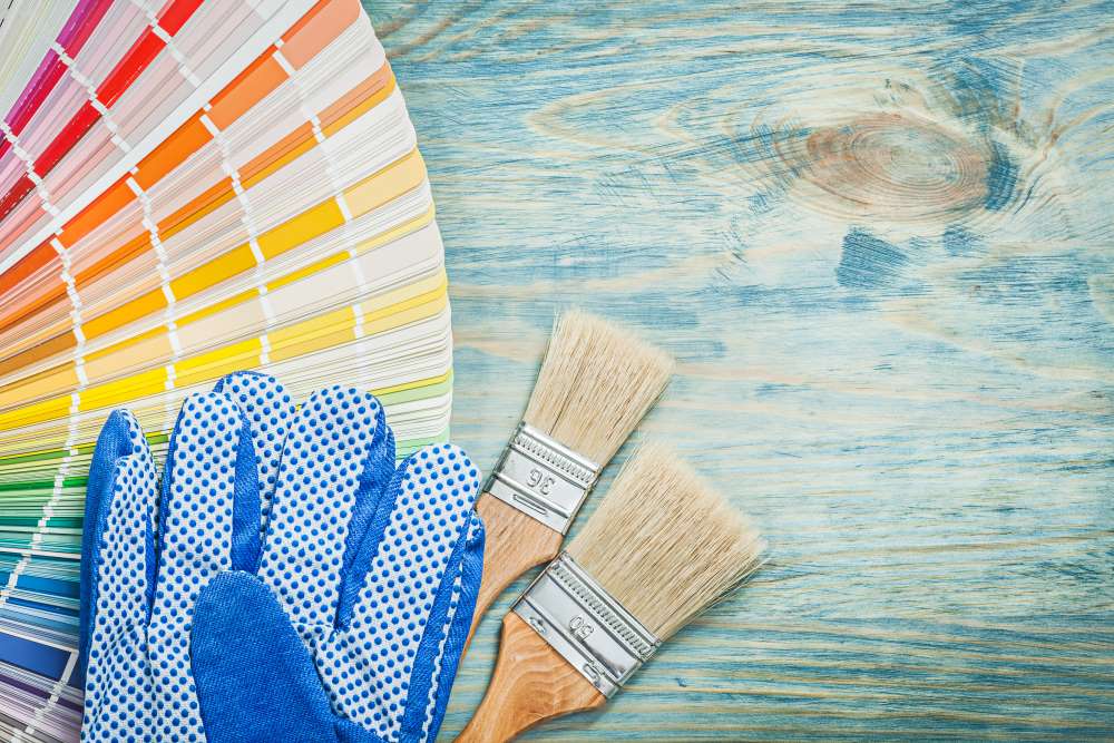 Painters' gloves, a paintbrush, and a wheel of color options lie on the ground as an investor plans to renovate their fix and flip investment.