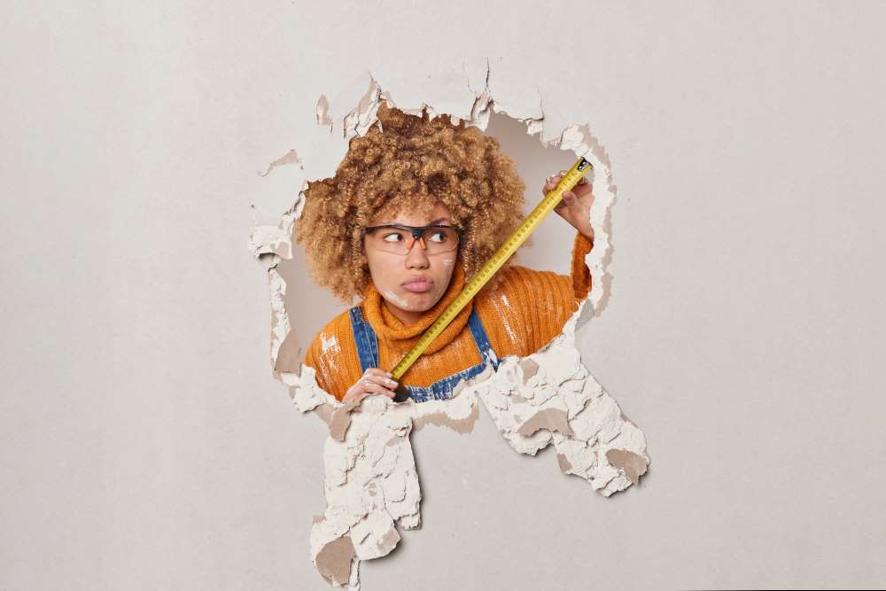 A woman measures a hole in the wall that needs repair in the house she intends to fix and flip with her Real Estate IRA. 