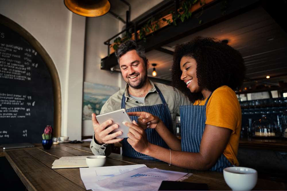 Small business owners looking at a tablet and smiling in their coffee shop business, to show that May is Small Business Month and that you can invest in a private business with a Self-Directed IRA.